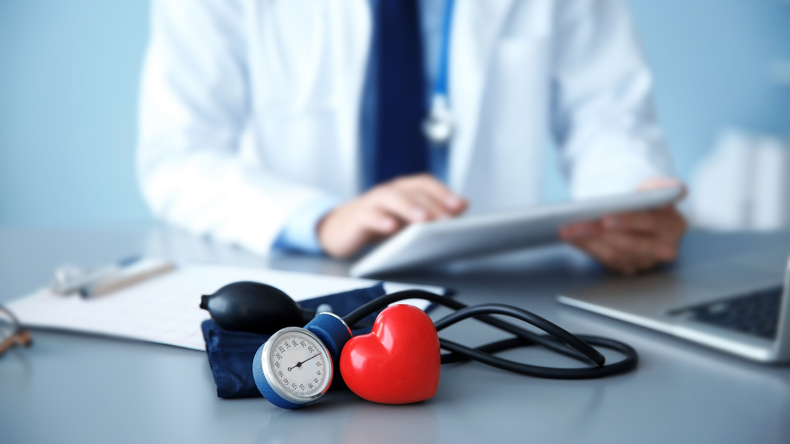 Médico trabajando en un escritorio con un monitor de presión arterial y un modelo de corazón sobre la mesa.