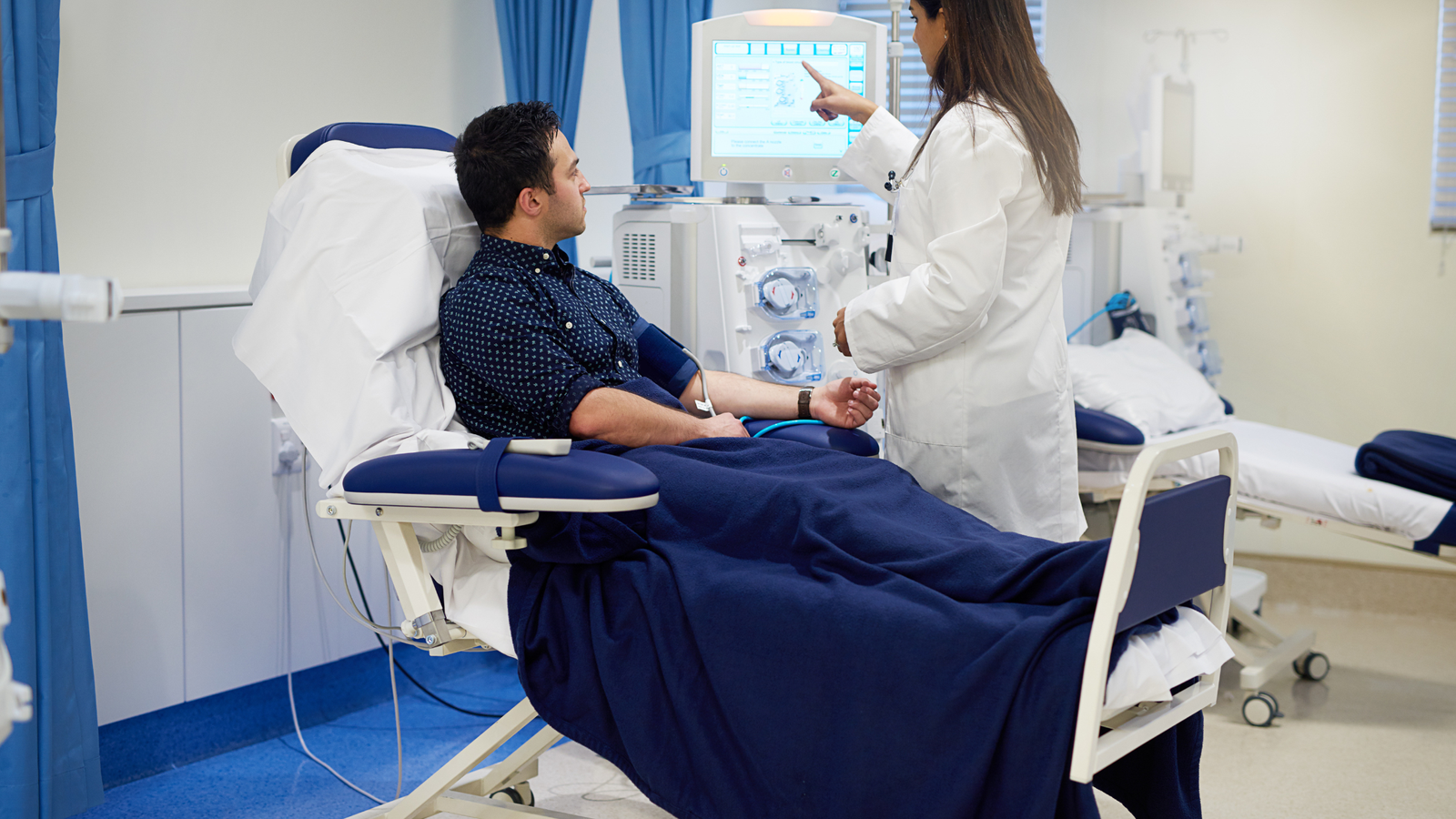 Muestra a un paciente en un ambiente médico especializado, posiblemente una clínica de hemodiálisis. El paciente está conectado a una máquina de diálisis, y un médico o enfermero parece estar explicando o ajustando el procedimiento.